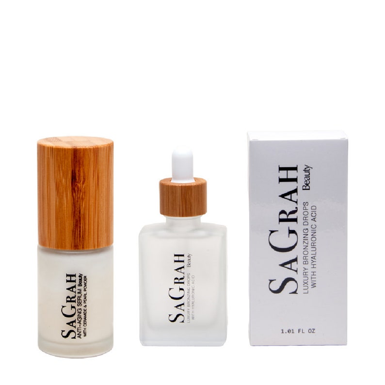 K&ouml;nnte beinhalten: Zwei Glasflaschen mit Bambusdeckeln und wei&szlig;en Etiketten. Die Flaschen enthalten Hautpflegeprodukte. Die Etiketten lauten "Sagrah Beauty" und "Luxury Bronzing Drops with Hyaluronic Acid". Die Flaschen stehen auf einem wei&szlig;en Hintergrund.