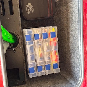 May include: Three clear plastic tackle boxes with blue latches, filled with fishing lures, are stacked inside a red carpeted storage compartment. The compartment has a black panel with the Bass Pro Shops logo.