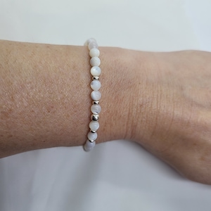 May include: A white beaded bracelet with silver accents. The bracelet is made of round beads and is worn on a person's wrist.