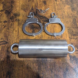 May include: A set of silver-colored handcuffs and a cylindrical metal object with ring handles. Two keys are also present. The items are arranged on a dark wooden surface, creating a contrast with the metallic objects.