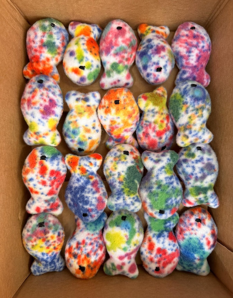 May include: A box filled with 20 colorful tie-dye fish plush toys. The fish are all white with different colored spots and swirls.