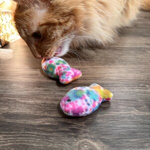 May include: A ginger cat playing with two colorful fish-shaped cat toys on a wood floor. The toys are made of soft fabric and have a tie-dye pattern.