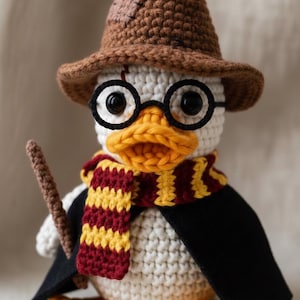 Può includere: Un'anatra bianca all'uncinetto con un cappello da mago marrone, occhiali rotondi neri e una sciarpa a righe rosse e oro. L'anatra ha anche un mantello nero e tiene in mano una bacchetta marrone. L'anatra ha zampe gialle e un becco giallo e arancione.