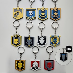 Könnte beinhalten: Eine Sammlung von Schlüsselanhängern mit verschiedenen Schilddesigns. Jeder Schlüsselanhänger hat einen Metallring und eine Kette mit einem Gummi- oder Kunststoffemblem in Farben wie Gelb, Blau, Schwarz, Rot und Weiß. Die Embleme zeigen Schädel- und Sternmotive.
