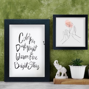May include: Two framed art prints with black frames hang on a green wall. One print has the words "Cold Air Dark Night Warm Fire Bright Stars" in black script. The other print features a line drawing of hands holding a flower. A white elephant statue and a potted plant are on a wooden shelf.