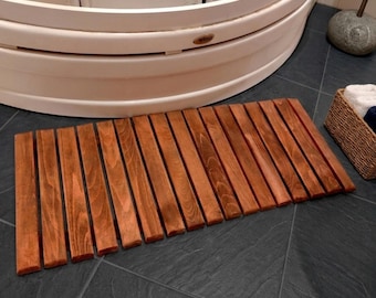 Könnte beinhalten: Ein rechteckiger Badteppich aus Holz mit vertikalen Lamellen. Der Teppich hat eine warme braune Farbe und liegt auf einem dunkelgrauen Fliesenboden. Eine weiße Badewanne und ein Weidenkorb mit Handtüchern befinden sich im Hintergrund.