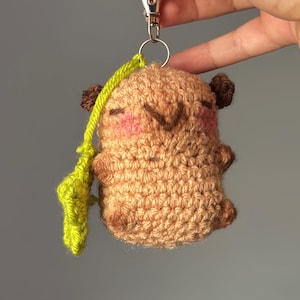 May include: A handmade crochet keychain featuring a small, brown creature with dark brown ears and rosy cheeks. A green leaf-shaped accent hangs from the keychain. The keychain is attached to a silver clasp.
