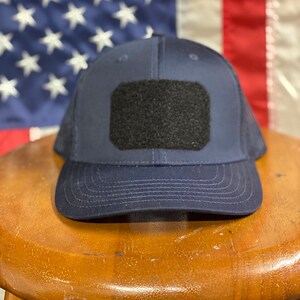 Pode incluir: Um bon&eacute; de caminhoneiro de malha azul marinho com um remendo de velcro preto na frente. O bon&eacute; est&aacute; sentado em um banquinho de madeira.