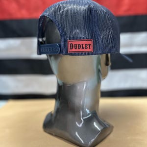 May include: Navy blue trucker hat with a mesh back and a curved peak. The hat has a red rectangular patch with the name "DUDLEY" in white block letters. The hat is displayed on a mannequin head.