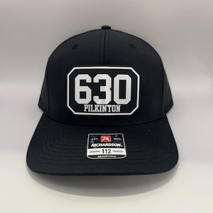 Pode incluir: Boné de caminhoneiro preto com um patch retangular branco com o número "630" acima da palavra "PILKINTON" em negrito. O boné tem uma aba curva e uma parte traseira em malha. A etiqueta da marca Richardson é visível.