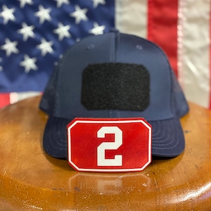 Op de afbeelding: Een donkerblauwe mesh trucker cap met een zwart klittenband patch en een rood patch met het nummer "2" in wit.