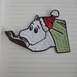 May include: Embroidered patch featuring a white cartoon character wearing a red Santa hat and reading a book. The patch has a green background and black border, perfect for adding to clothing or accessories.