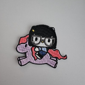 Könnte beinhalten: Gestickter Aufnäher mit einer Cartoonfigur mit schwarzen Haaren, Brille und blauem Hemd, die ein lila Einhorn reitet. Das Einhorn hat eine rote Mähne und einen roten Schweif. Für Kleidung oder Accessoires.