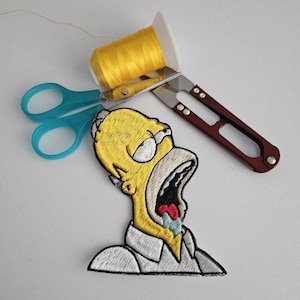 May include: Embroidered patch featuring Homer Simpson with his mouth open in a scream. The patch is yellow, white, red, and blue. Also pictured are a spool of yellow thread, blue scissors, and a brown and silver craft knife.