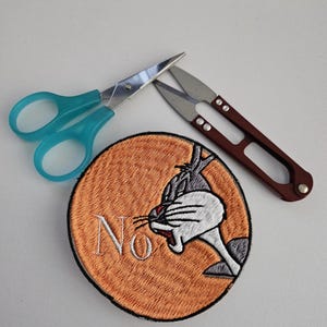 May include: An orange Bugs Bunny patch with the word "No" embroidered in white, next to a pair of teal scissors and a pair of brown thread snips. The patch features Bugs Bunny's face in black, white, and gray.