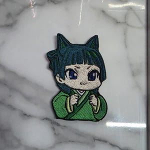 May include: Embroidered patch depicting a cartoon character with teal hair and cat-like ears. The character is dressed in a green robe with cream trim and has blue eyes. The patch is designed for application on clothing or accessories.
