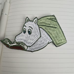 May include: Embroidered patch featuring a white, cartoon-style creature with large eyes, reading a book. The creature has a green cape. The patch is detailed with various colors and textures, suitable for application on clothing or accessories.