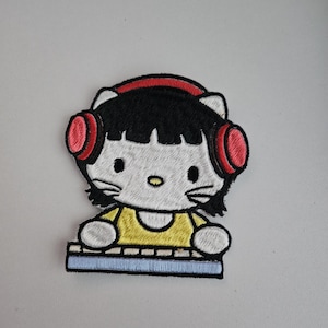 Pode incluir: Patch bordado com um gato de desenho animado com cabelo preto, usando fones de ouvido vermelhos. O gato está vestido com uma camisa amarela e está posicionado em frente a um teclado azul. O patch é branco, preto, vermelho, amarelo e azul.