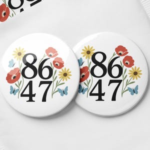 Puede incluir: Un conjunto de botones redondos blancos con los números "86 47" en negro, rodeados de amapolas rojas, margaritas amarillas y mariposas azules. Los botones están empaquetados en una funda de papel blanco.