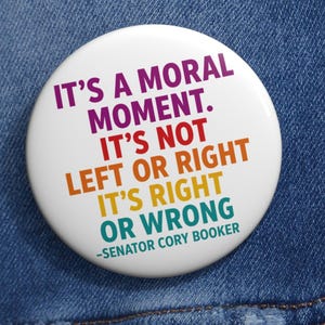 May include: A white button badge with the text "IT'S A MORAL MOMENT. IT'S NOT LEFT OR RIGHT IT'S RIGHT OR WRONG -SENATOR CORY BOOKER" in purple, red, orange, yellow, and green. The badge is set against a blue denim jacket.