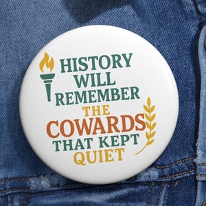 May include: A round white pin-back button featuring a torch icon and a wheat stalk graphic. The text reads: HISTORY WILL REMEMBER THE COWARDS THAT KEPT QUIET. The lettering uses a mix of dark green, orange, and yellow colours on a white background.