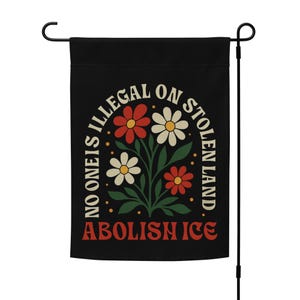May include: Black garden flag with the text "NO ONE IS ILLEGAL ON STOLEN LAND" and "ABOLISH ICE" in white and red. A floral design with red, white, and yellow flowers is in the center. The flag is attached to a black metal pole.