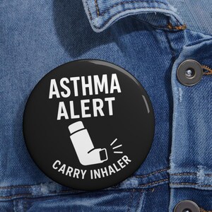 May include: A round black pin-back button featuring white text that reads ASTHMA ALERT and CARRY INHALER, with a white graphic of an inhaler positioned between the two lines of text. The button is attached to a blue denim fabric surface.