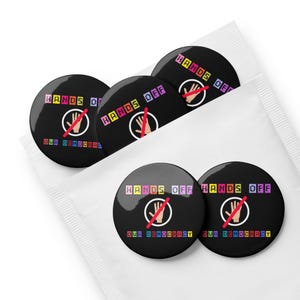 Puede incluir: Un conjunto de cinco botones redondos negros y blancos con el texto "Hands Off Our Democracy" en letras de bloque de colores. Cada botón presenta una mano roja con una línea diagonal que la atraviesa.