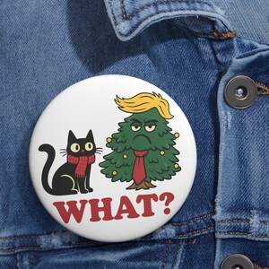 Puede incluir: Un botón blanco con un gato negro con una bufanda roja y un árbol de Navidad gruñón con un tupé amarillo y una corbata roja. La palabra "WHAT?" está impresa en rojo debajo de las ilustraciones.