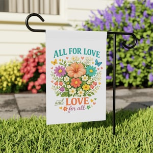 May include: A white garden flag hanging on a black metal stand, featuring a colourful floral bouquet with butterflies and hearts. The text reads ALL FOR LOVE in teal and and LOVE FOR ALL in orange, surrounded by pink, yellow, and blue flowers.