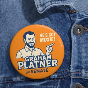 Peut inclure: Un bouton rond orange avec le texte "HE'S GOT MOXIE!" et "GRAHAM PLATNER for SENATE" en bleu et blanc. Le bouton présente une illustration de dessin animé d'un homme pointant vers la droite.