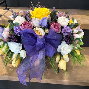 May include: A colourful floral display with a large purple bow. The arrangement includes yellow roses, white and pink roses, purple and white tulips, and various other flowers and foliage. The flowers are arranged in a semi-circular shape.