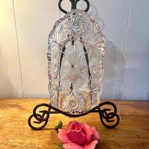 May include: A clear, cut-glass dish with starburst and floral patterns, displayed on a black wrought-iron stand. A pink ceramic rose with green leaves rests in front of the stand, creating a decorative display.