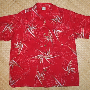 May include: Red short-sleeved button-down shirt with a tropical floral pattern. The shirt features a vibrant red background with white and gold bird-of-paradise flowers and leaf designs. The shirt has a classic collar and gold buttons.