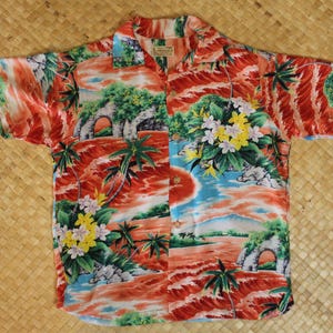 Puede incluir: Camisa de manga corta con estampado tropical. El diseño presenta palmeras, flores y arcos en tonos rojos, naranjas, verdes y azules. La camisa abotonada está colocada sobre una superficie tejida.