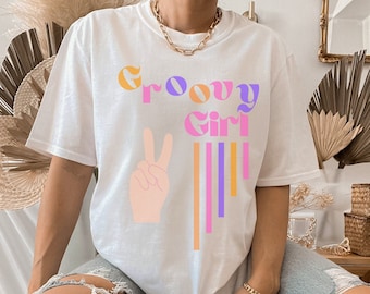 Könnte beinhalten: Weißes T-Shirt mit dem Text "Groovy Girl" in bunten Buchstaben und einer Peace-Sign-Grafik. Das Shirt wird von einer Person mit einer goldenen Kettenkette getragen.