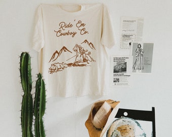 May include: A beige t-shirt with a brown graphic of a cowboy riding a horse and the text "Ride 'Em Cowboy Co."