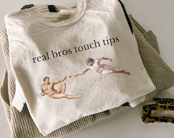 May include: A beige t-shirt with the text "real bros touch tips" printed on the front. The shirt features a graphic of two figures reaching towards each other.