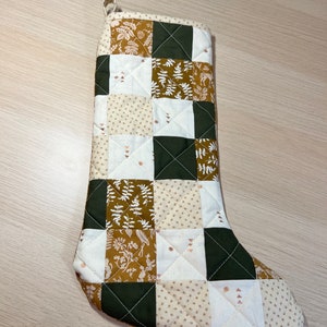 May include: A quilted Christmas stocking with a patchwork design. The stocking features squares of white, olive green, and tan fabric with floral and fern patterns. A loop is attached at the top for hanging. The stocking is approximately 18 inches long.