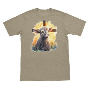 May include: A beige t-shirt with a watercolor illustration of a lamb standing in front of a wooden cross. The lamb is looking up at the cross, and the background is a soft yellow and orange.