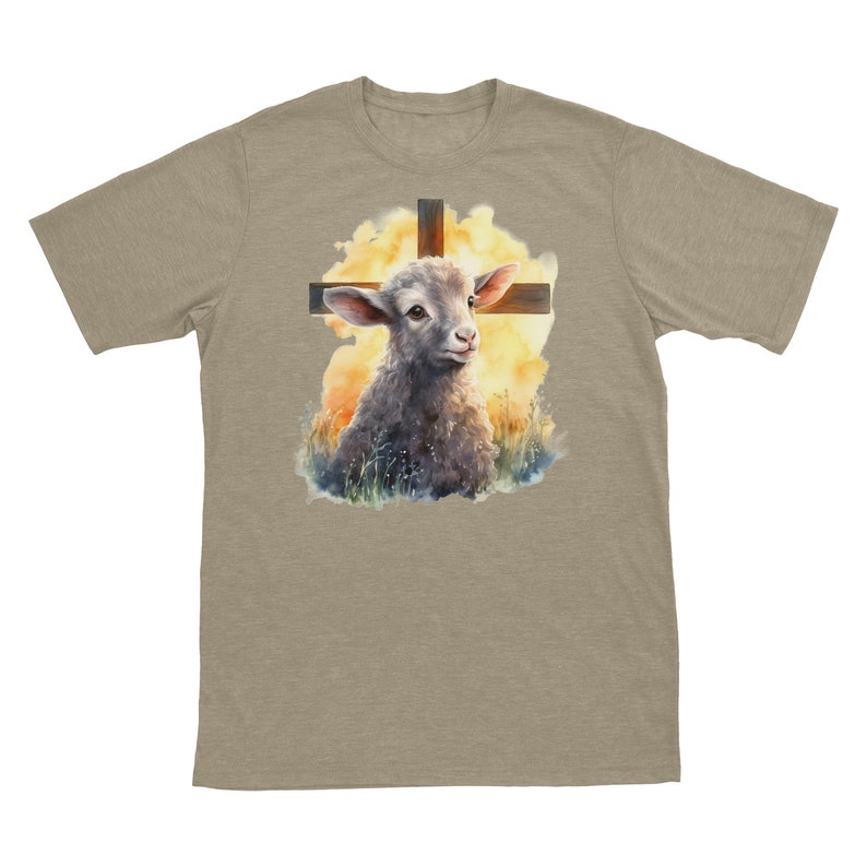 May include: A beige t-shirt with a watercolor illustration of a lamb standing in front of a wooden cross. The lamb is looking up at the cross, and the background is a soft yellow and orange.