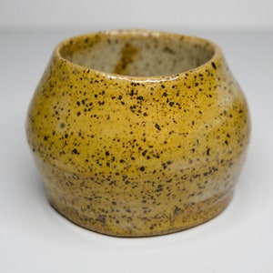 Può includere: Una piccola ciotola in ceramica gialla con una glassa marrone macchiata. La ciotola ha una forma arrotondata e un bordo leggermente svasato.