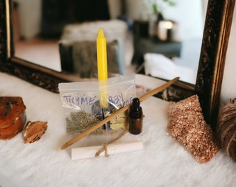 May include: A white surface with a yellow candle, two small bags labeled "THYME" and "BAY", a wooden stick, a small brown bottle with a dropper, a rolled up piece of paper tied with twine, and a brown ball of yarn.