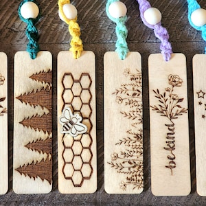 Puede incluir: Seis marcapáginas de madera con diferentes diseños, incluyendo flores, árboles, un panal con una abeja y estrellas. Cada marcapáginas tiene un borla de colores.