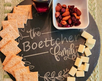 Puede incluir: Una tabla de servir de pizarra negra con el texto "the Boettcher family" inscrito en ella. La tabla está decorada con un diseño de hojas y presenta una variedad de bocadillos, que incluyen galletas, cubos de queso, arándanos secos y almendras.