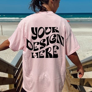 May include: A pink t-shirt with the text "YOUR DESIGN HERE" printed in black on the back. The shirt is being worn by a person standing on a wooden staircase with a beach in the background.