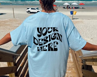 May include: A person wearing a light blue t-shirt with the text "YOUR DESIGN HERE" in black on the back. The person is standing on a wooden staircase with a beach in the background.