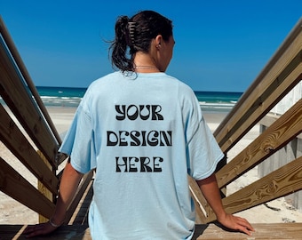 May include: A person wearing a light blue t-shirt with the text "YOUR DESIGN HERE" printed on the back. The person is sitting on a wooden staircase with a beach in the background.