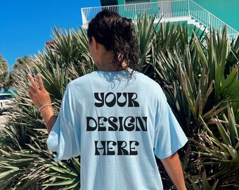 May include: Light blue oversized t-shirt with the text "YOUR DESIGN HERE" in black, set against a backdrop of lush green foliage and a turquoise building under a clear blue sky. The shirt is a relaxed fit.