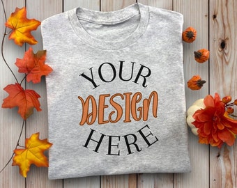 May include: A gray t-shirt with the text "YOUR DESIGN HERE" in black and orange lettering. The shirt is folded and laid on a wooden surface with fall-themed decorations.